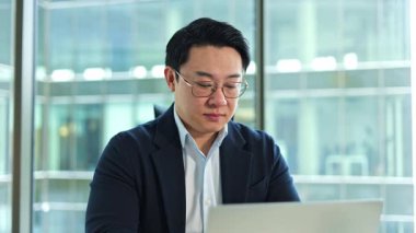 Modern ofiste dizüstü bilgisayarla çalışan Koreli iş adamı. İşyerindeki bilgisayar klavyesinde yazmakla meşgul. Masada not defteri kullanan, gözlüklü, kendine güvenen bir girişimci..