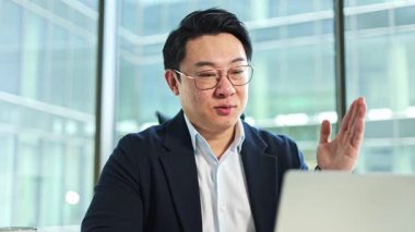 Modern ofiste kendine güvenen Koreli iş adamı bilgisayarını kullanıyor. Yakışıklı Asyalı erkek iş ortaklarıyla çevrimiçi konferans yapıyor..