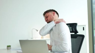 Modern ofiste dizüstü bilgisayarla uzun süre çalıştıktan sonra yorgun işadamı omzuna ağrıdan dolayı masaj yapıyor. Erkek çalışan boynunu uzatıyor..