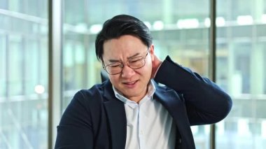 Stresli Koreli iş adamının modern bir ofiste boyun ağrısı var. Yorgun Asyalı adam boynuna masaj yapıyor. Gözlüklü erkek girişimci iş yerinde kendini rahatsız hissediyor.