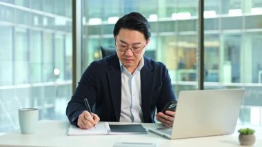 Akıllı telefonları kontrol ederken Koreli işadamı defterine notlar yazıyor. Modern ofiste masa başında çalışan yetişkin bir girişimci..