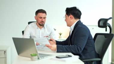 İki işadamı modern bir ofiste, bir masada dizüstü bilgisayarla evrakları gözden geçiriyorlar. Bir adam meslektaşına bazı detayları açıklıyor..