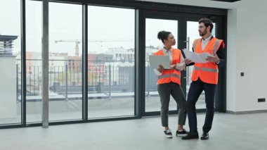 Güvenlik yelekleri giyen iki profesyonel mimar planları kontrol ediyor ve boş bir ofis binasında bir projeyi tartışıyorlar..