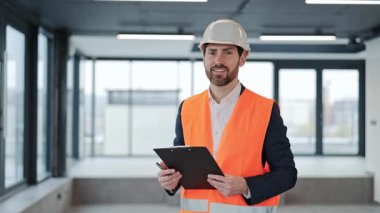 Yeni modern binanın içinde resmi kostüm, kask ve güvenlik yeleği konusunda başarılı bir mühendis duruyor. Erkek yatırımcı erkek mimar pano tutuyor, gülümsüyor ve doğrudan kameraya bakıyor..