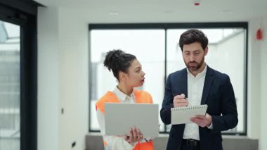 Profesyonel yatırımcı ve inşaat uzmanı boş bir bina içinde bir projeyi tartışıyor. Yatırımcı not defterine, turuncu yelek uzmanı dizüstü bilgisayar tutuyor..