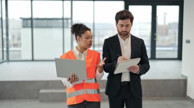Profesyonel yatırımcı ve mühendis modern ofiste inşaat planlarını tartışıyor. Yatırımcı notlar alır, mühendis dizüstü bilgisayar tutar. Takım çalışması ve başarı.