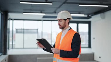 Turuncu yelek ve beyaz kasklı başarılı bir mühendis elinde bir panoyla parlak geniş bir ofiste duruyor ve kameraya bakıyor..