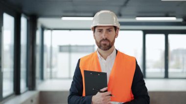 Kendine güvenen mühendis, inşaat halindeki bir binanın içinde elinde bir pano tutarak gülümsüyor. Emniyet yeleği ve şapka takıyor..
