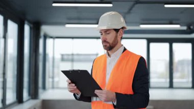 Turuncu güvenlik yeleği ve kask takan başarılı mühendis inşaat alanındaki çalışmalarını kontrol ediyor ve gülümsüyor..
