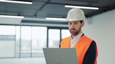 Turuncu yelek ve beyaz kask giyen başarılı bir mühendis boş bir ofis binasında dizüstü bilgisayarda çalışıyor. Mutlulukla gülümsüyor..