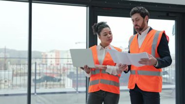 Güvenlik yelekleri giyen iki profesyonel mimar proje planlarını gözden geçirir ve bir inşaat alanında dizüstü bilgisayar kullanarak proje detaylarını tartışır..