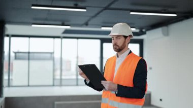 Resmi kıyafet ve yelek mühendisi ofis binasını denetliyor, kontrol listesi tutuyor. İnşaat, teftiş ve geliştirme.
