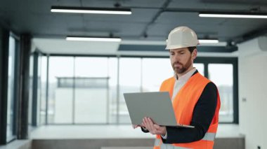 Ofiste çalışan başarılı turuncu yelek ve beyaz kasklı mühendisin portresi. Dizüstü bilgisayar kullanan profesyonel bir uzman. Bilgisayarla kendine güvenen bir iş adamı.