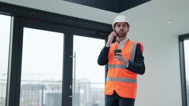 Resmi kıyafet ve güvenlik yeleği içinde başarılı bir mühendis kahve içiyor ve modern bir ofis binasında akıllı telefonuyla konuşuyor..