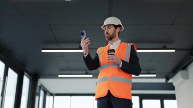 Ofis binasında başarılı bir mühendis takım elbise ve güvenlik yeleği giyiyor kahve ve telefonda görüntülü sohbetlerden hoşlanıyor.