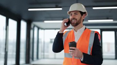 Resmi giyim ve şapkalı mutlu mühendis akıllı telefondan konuşuyor ve şantiyede kahve tutuyor. Neşeli ve kendinden emin.