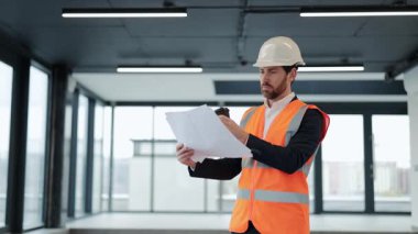 Resmi giyimde ve güvenlik yeleğinde başarılı bir mimar planları gözden geçirir ve modern ofis alanında kahve içer. Kendini odaklanmış ve kararlı hissediyor..