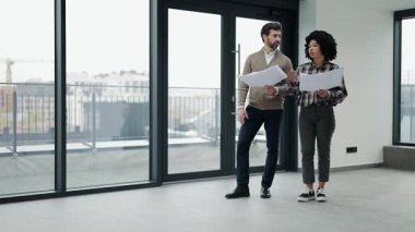 Gündelik giysiler içindeki profesyonel meslektaşlar şehir manzaralı modern ofis alanının planlarını tartışırlar. İşbirliği ve coşku ortada..