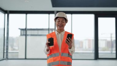 Kendine güvenen mühendis modern ofiste telefonda kahve içip görüntülü sohbet ediyor. Şapkalı ve üniformalı profesyonel işçi kendini mutlu hissediyor..