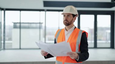 Resmi kıyafet ve güvenlik yeleği giyen profesyonel mimar modern ofis alanının planlarını inceliyor. Projenin detaylarını düşünüyor..