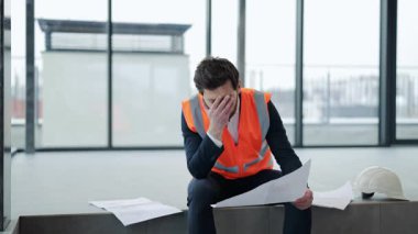 Takım elbiseli ve güvenlik yeleği giyen stresli mimar modern ofis binasının planlarını inceliyor. Hayal kırıklığına uğramış ve hayal kırıklığına uğramış hissediyor..