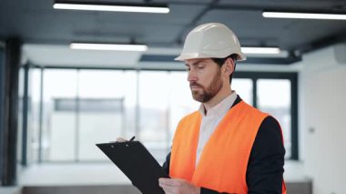 Turuncu yelek giymiş profesyonel bir mühendisin portresi ve bir not panosuna not alıyor. O, inşaat halinde olan modern bir binada duruyor. Ciddi bir ruh hali..