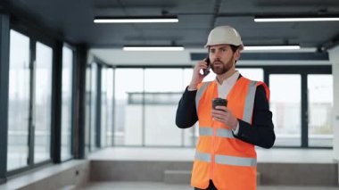 Turuncu yelek ve kask giyen başarılı bir mühendis telefonla konuşuyor ve kahve içiyor. Yeni ofiste kendini mutlu ve memnun hissediyor..