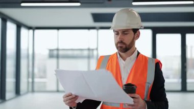 Resmi takım elbise ve güvenlik yeleği içinde başarılı bir mimar planları gözden geçirir ve modern ofiste kahve içer. Odaklanmış hissediyor..