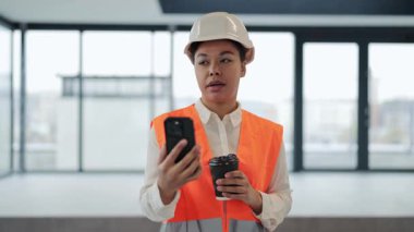 Başarılı turuncu yelek ve beyaz şapka giyen kadın mühendis kahve içerek görüntülü konuşma yapıyor ve modern ofiste telefon kullanıyor. Mutlu yüz, iyi ruh hali.