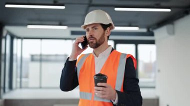 İnşaat yeleği, kostüm ve kaskta başarılı bir mühendis, kahve içiyor, akıllı telefon kullanıyor ve ofiste kendini mutlu hissediyor..