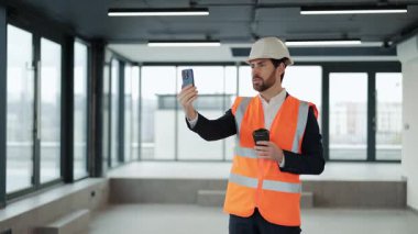 Resmi aşınma ve güvenlik yeleğinde başarılı bir mühendis. Panoramik pencereli boş ofis alanındaki akıllı telefondan video çağrısı alıyor..