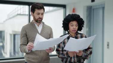 Odaklanmış meslektaşlar proje detaylarını modern bir ofiste tartışırlar. Profesyonel kadın ve erkek şık kıyafetler giyiyor. Takım çalışmasını ve uzmanlığını gösteren belgeleri gözden geçiriyorlar..