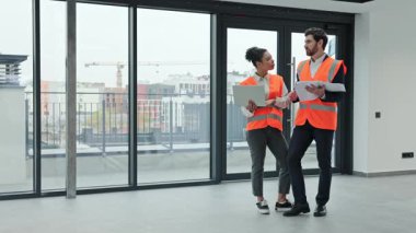 Kadın profesyonel mühendis ve yatırımcı boş bir ofis binasının planlarını tartışıyor. Kendilerinden emin ve işbirlikçi hissediyorlar, şık günlük kıyafetler ve güvenlik yelekleri giyiyorlar..