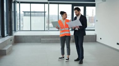 Profesyonel mimar ve yatırımcı modern bir ofisin planlarını tartışıyor. Resmi giyinmiş bir yatırımcı ve güvenlik yelekli günlük kıyafetler giymiş bir mimar..