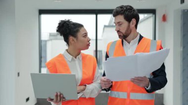 Başarılı çeşitli meslektaşlar modern bir binanın planlarını tartışıyor. Güvenlik yelekleri ve resmi kıyafetler giyen profesyonel mimarlar, dizüstü bilgisayar ve belge kullanan bir projede işbirliği yapıyorlar..