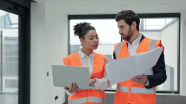Turuncu yelekli başarılı meslektaşlar inşaat alanının planlarını tartışıyor. Odaklanmış işçiler, proje başarısı ve verimliliğinin sağlanması için dizüstü bilgisayardaki bilgileri kontrol ediyorlar.