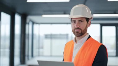 Bilgisayarlı başarılı mühendisin portresi modern ofis alanındaki kameraya koruyucu iş kıyafeti giyip gülümsüyor. İnşaat müfettişi mutlu..