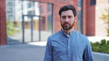 Yakışıklı bir iş adamı, şık ve rahat gömlekli. Başparmaklarını kaldırıyor ve gülümsüyor. Mutlu erkek dışarıda ofis binasının yanında duruyor. Pozitif duygu, uzlaşma ve başarı.