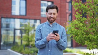 Ofis binasının yanında akıllı telefon kullanan sıradan giysili yakışıklı bir iş adamı. Başarılı bir adam gülümseyerek dışarıda mutlu olur. Kullanılan mobil aygıt.