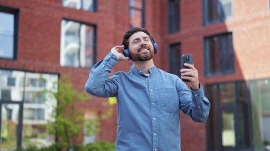 Gündelik giysiler içindeki olumlu adam kulaklıkla müzik dinlemekten hoşlanır, modern bir binanın önünde ritme ayak uydurur..