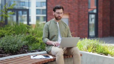 Açık havada dizüstü bilgisayar üzerinde çalışan odaklanmış bir iş adamı. Profesyonel erkek, bankta oturuyor ve dizüstü bilgisayar kullanıyor. Şehir yeşili. İş ve teknoloji kavramı.