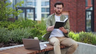 Günlük giysiler içinde yakışıklı bir iş adamı, elinde defter var ve dışarıda video görüşmesi yapıyor. Ciddi ve odaklanmış görünüyor, yanında bir dizüstü bilgisayarla..