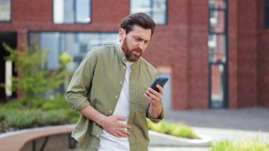 Adam dışarıda akıllı telefon kullanırken mide ağrısını hisseder. Sakallı adam akıllı telefon kullanırken karnını tutuyor. Günlük giysiler içinde mutsuz bir erkek..