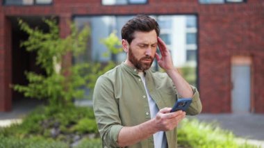 Sinirli iş adamı ofis yakınlarında telefonundan kötü haberler okurken baş ağrısı ve stres hissediyor. Sıradan giyinmiş..
