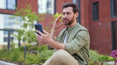 Yakışıklı, neşeli erkek serbest çalışanın olumlu duyguları var. Yakışıklı Adam Şehir Bahçesi 'nde cep telefonuyla video konferans sohbetinden hoşlanıyor