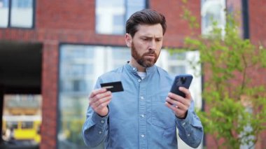 Kafası karışmış işadamı akıllı telefondan online ödeme yapmada sorun yaşıyor. Yakışıklı, sıradan giyinmiş, dışarıda dikilen sinirli ve sinirli bir erkek..