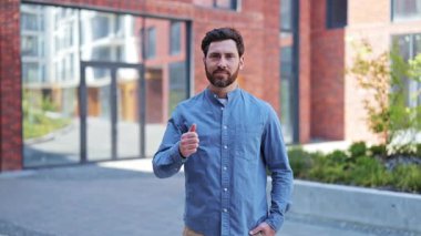 Modern ofis binasının yakınında, sıradan kıyafetler içindeki yakışıklı iş adamının portresi onaylıyor. Başarılı İşadamı Ofis Binası 'nın yakınında Başparmak Verdi. Erkek öğretmen neşeli ve kendinden emin hissediyor..