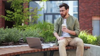 Günlük giysiler içinde yakışıklı bir iş adamı internette iş görüşmesi yapıyor ve dışarıda kahve içiyor..