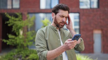 Sinirli iş adamı dışarıda telefonda kötü haberler okurken stresli hissediyor. Sıradan giyinmiş, hayal kırıklığı ve endişe yaşıyor..