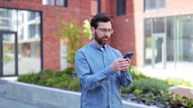 Ofisin yanındaki telefonu kullanarak gözlüklü ve günlük kıyafetler giyen yakışıklı bir iş adamı. Dışarıda gülümseyen neşeli iş adamı. İnternette başarılı bir iş adamı sohbeti.